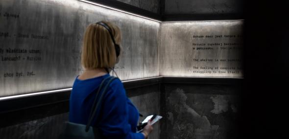 Kobieta stoi w bunkrze zrekonstruowanym na wystawie "Wokół nas morze ognia". Zwiedzająca ma słuchawki na uszach, a przed sobą trzyma audioprzewodnik. Na ścianach bunkra znajdują się zapiski ze wspomnień osób ukrywających się w getcie warszawskim.