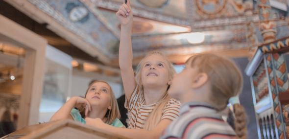 Trzy dziewczynki patrzą na sklepienie synagogi w Gwoźdźcu zrekonstruowane na wystawie stałej w POLIN. Jedna z dziewczynek trzyma ołówek w uniesionej ręce.