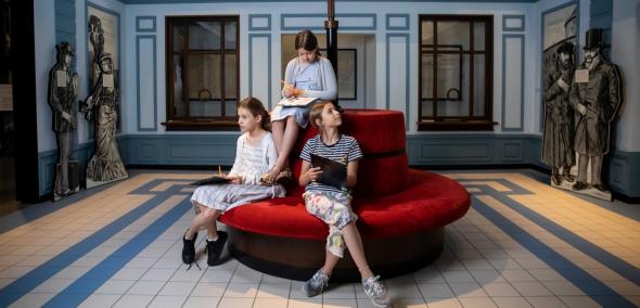 Trzy dziewczynki siedzą na kanapie w galerii "Wyzwania nowoczesności" na wystawie stałej w Muzeum POLIN. W dłoniach trzymają podkładki na kartki i ołówki.