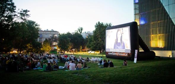 Widzowie siedzą na łące i oglądają film prezentowany na ekranie rozstawionym przy budynku Muzeum POLIN.