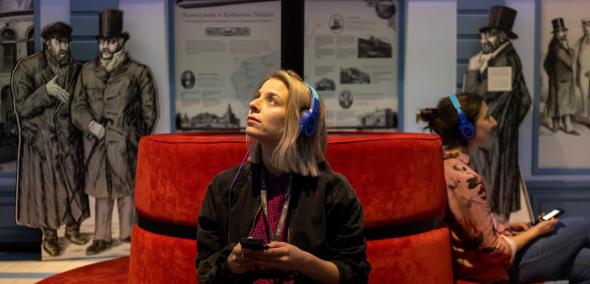 Dwie kobiety siedzą na czerwonej kanapie w galerii "Wyzwania nowoczesności" na wystawie stałej w Muzeum POLIN. Obie mają słuchawki na uszach i trzymają w dłoniach audioprzewodniki.