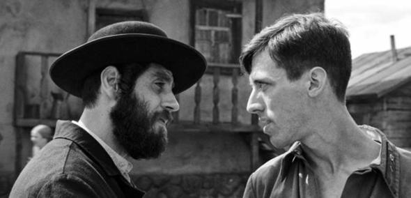 Czarno-biały kadr z filmu "Shttl" - mężczyzna z ciemną brodą i w kapeluszu celuje palcem w pierś drugiego mężczyzny.
