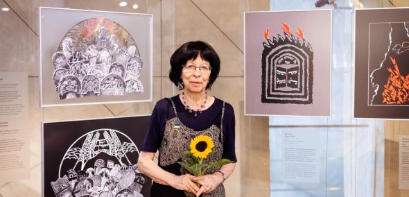 Monika Krajewska trzyma w dłoniach kwiat słonecznika. Za nią na szklanych ściankach znajdują się jej prace z cyklu "Płonące"