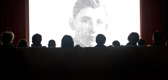 Grupa ludzi ogląda czarnobiały film. Kadr filmu przedstawia młodego mężczyznę patrzącego na wprost.