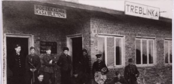 Czarno-białe zdjęcia przedstawia grupę ciepło ubranych ludzi, przed budynkiem dawnego dworca kolejowego w Treblince
