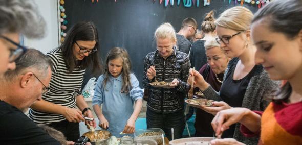 Międzypokoleniowe warsztaty kulinarne w JCC w ramach TISZ Festiwalu Żydowskiego Jedzenia - Muzeum POLIN, na zdjęciu widać młodych i dorosłych stojących wokół kuchennego stołu i zajętych przygotowywaniem potraw. W tle czarna tablica z kolorowymi chorągiewkami
