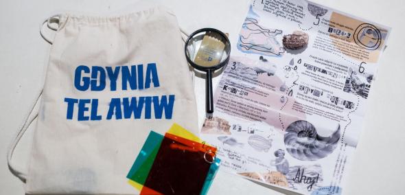 Rodzinna ścieżka zwiedzania wystawy "Gdynia - Tel Awiw" w Muzeum POLIN. Na zdjęciu: Plecak podróżnika: biały plecak-worek z niebieskim napisem "Gdynia - Tel Awiw", kolorowe folie, lupa, plan detektywa