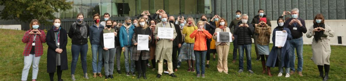 Grupa osób w jesiennych ubraniach stoi przed budynkiem Muzeum POLIN. Część trzyma transparenty, część dłonie złożone w symbol serca. Apel ws. kryzysu uchodźczego na granicy Polski i Białorusi