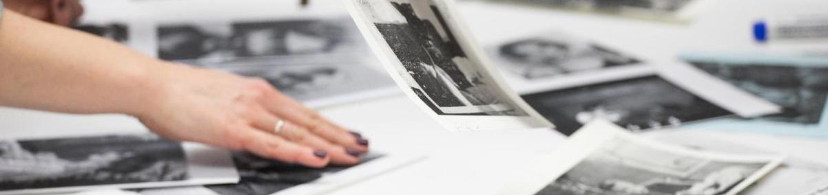 Czarno-białe zdjęcia leżą na stole. Jedna osoba dotyka fotografię, druga podnosi jedno zdjęcie.