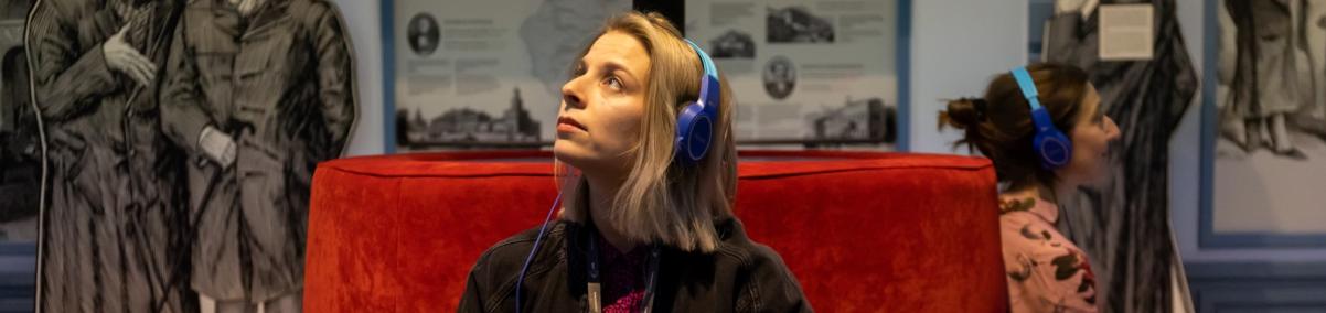 Dwie kobiety siedzą na czerwonej kanapie w galerii "Wyzwania nowoczesności" na wystawie stałej w Muzeum POLIN. Obie mają słuchawki na uszach i trzymają w dłoniach audioprzewodniki.