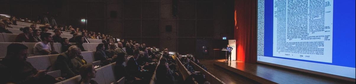 Uczestnicy wykładu siedzą słuchając prowadzącego spotkanie i oglądają pokaz slajdów w sali audytorium Muzeum Polin