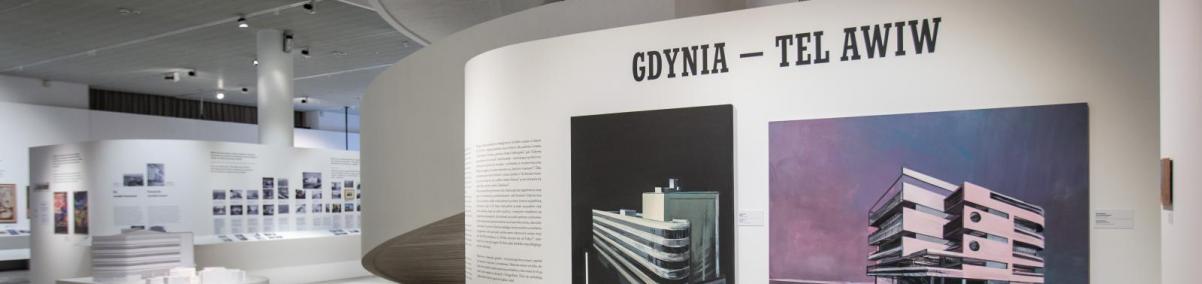 Poranne ptaszki - oprowadzanie po wystawie "Gdynia - Tel Awiw" dla osób ze spektrum autyzmu w Muzeum POLIN. Na zdjęciu przestrzeń wystawy, podzielona białymi ścianami działowymi, na pierwszym planie ściana z tytułem wystawy i dwoma obrazami w kolorystyce czarnej, fioletowej, przedstawiające budynki modernistyczne