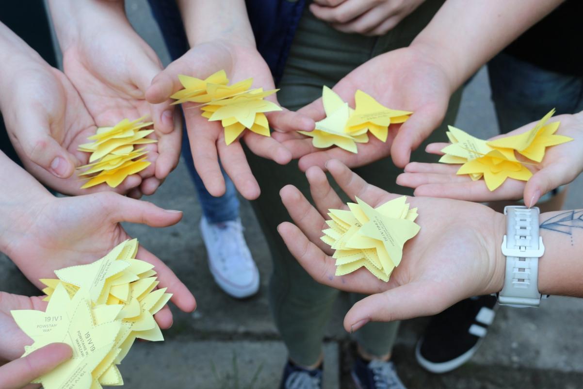 Na zdjęciu widać osiem dłoni, na których leżą żółte papierowe żonkile, symbol akcji społeczno-edukacyjnej Żonkile