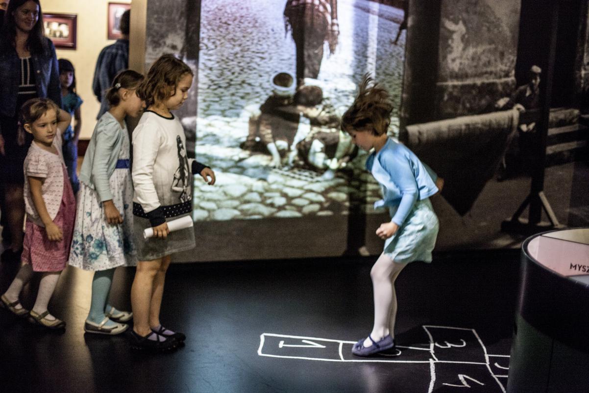 Czworo dzieci gra w klasy, w siatce wyrysowanej na podłodze w jednej z części wystawy stałej Muzeum POLIN