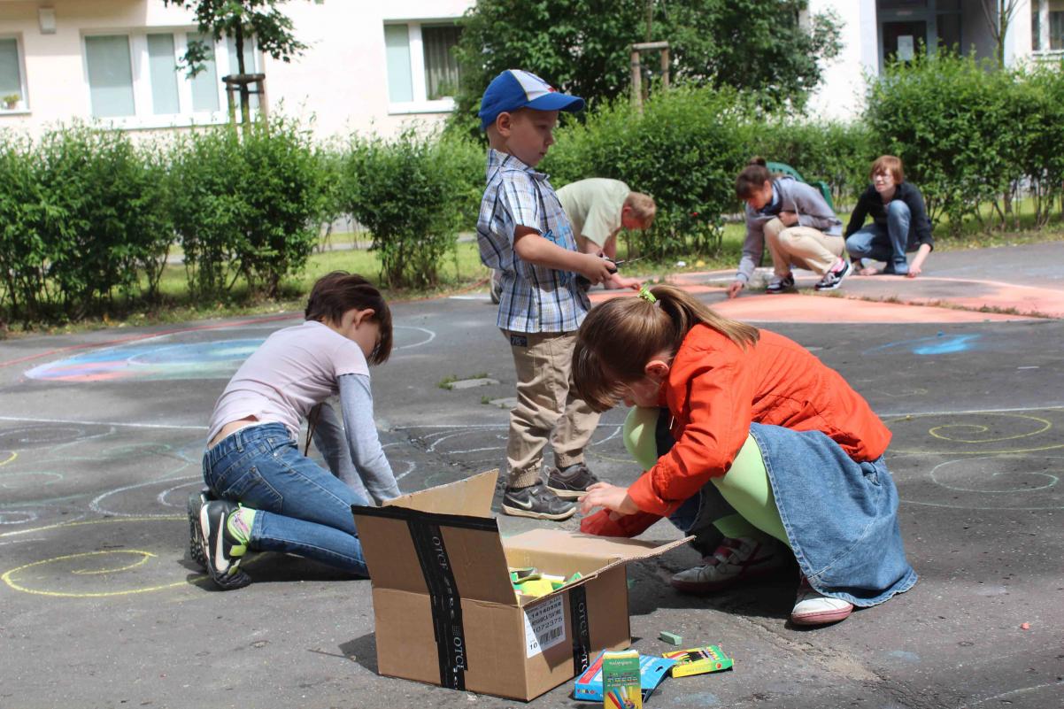 Dzieci bawią się na podwórku wśród budynków. Na asfaltowej nawierzchni malują kolorowymi kredami