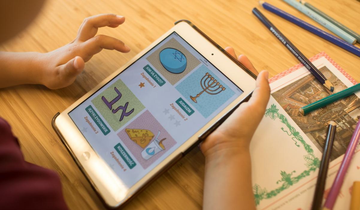 Widać ręce dziecka, które trzymają tablet otwarty na stronie z quizami dla dzieci "Żydowski, co to znaczy?"