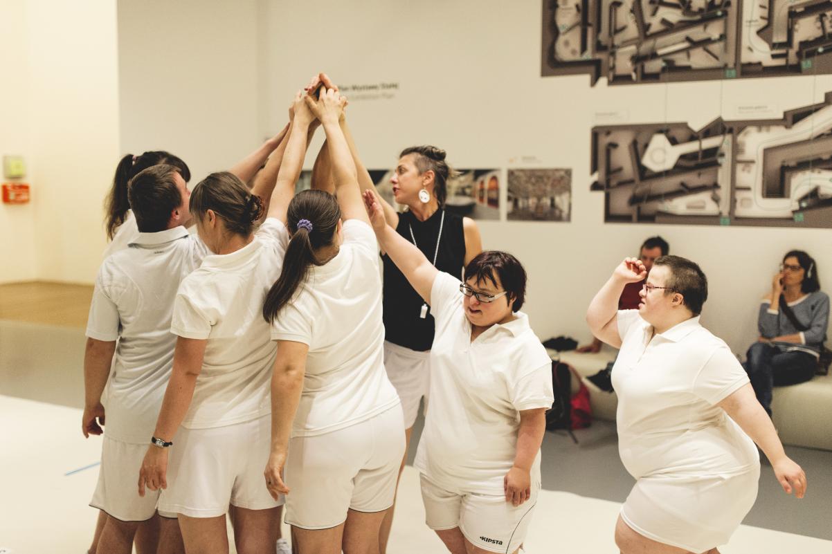 Grupa młodych osób ubranych w białe stroje sportowe gromadzi się na środku sali, każdy wznosi rękę jak do zbiórki popularnej zabawy "palec pod budkę".