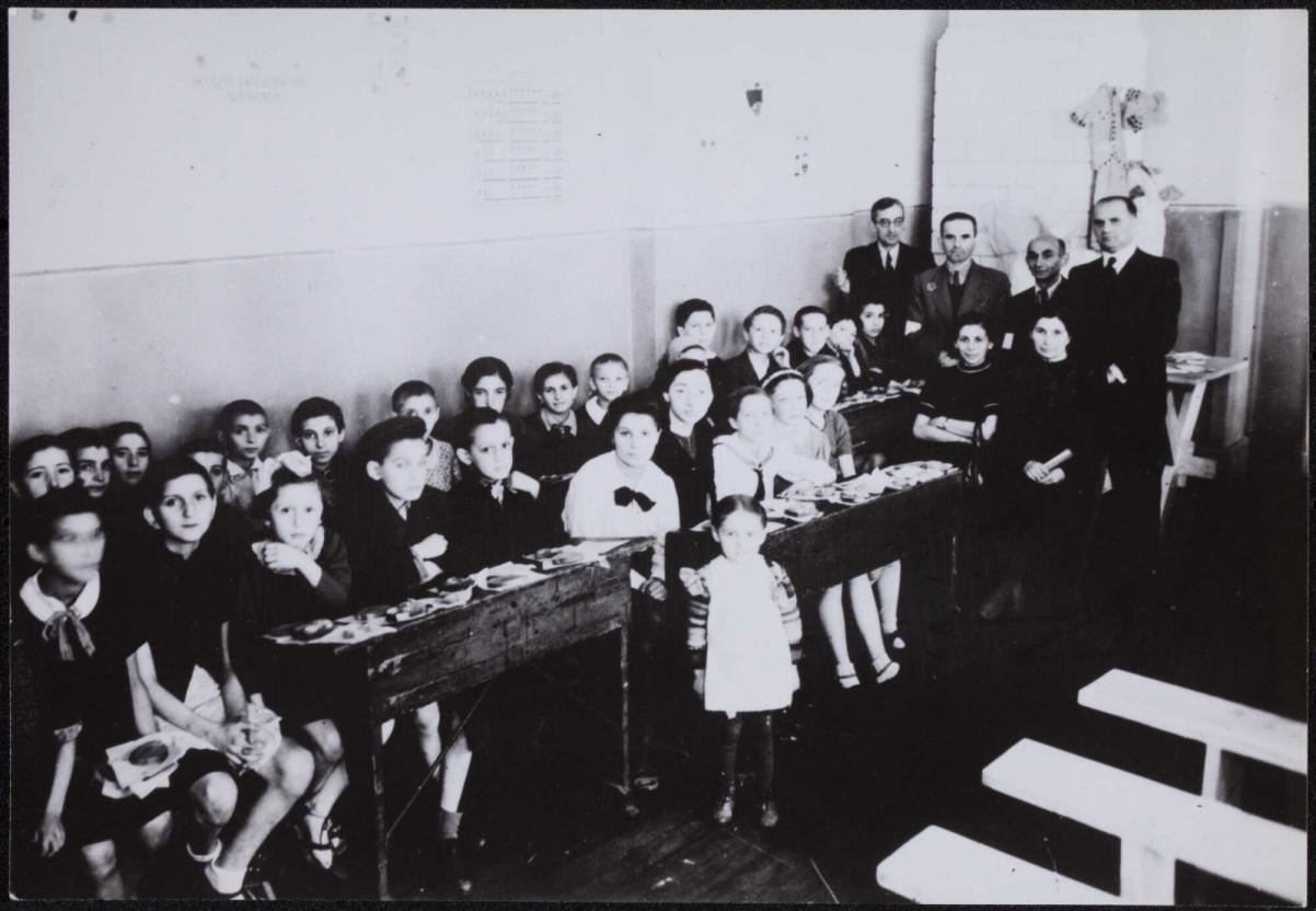 Zdjęcie przedstawia klasę szkolną. W ławkach siedzą dzieci, za nimi w głębi stoi grupa nauczycielek i nauczycieli. Zdjęcie czarno-białe.