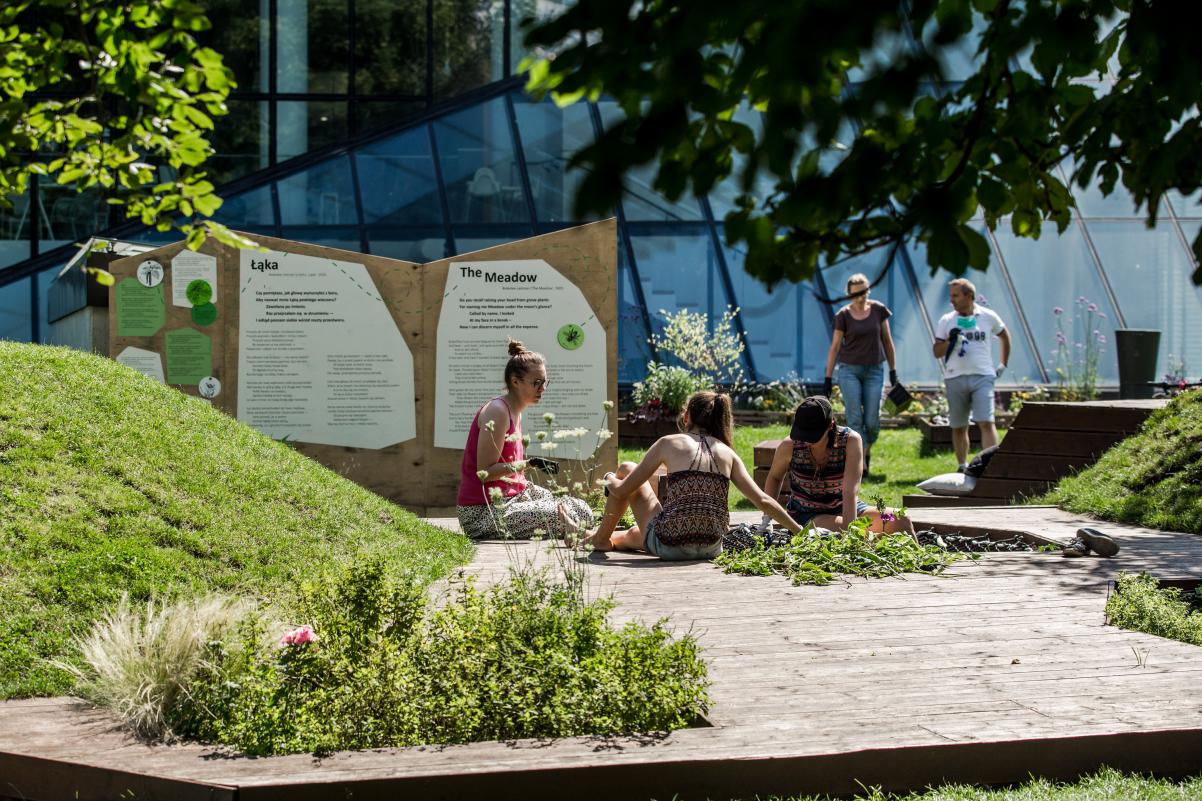 Na obrazie widzimy grupę osób - kobiet i mężczyzn, siedzących na łące obok muzeum, podczas letniego, słonecznego dnia.