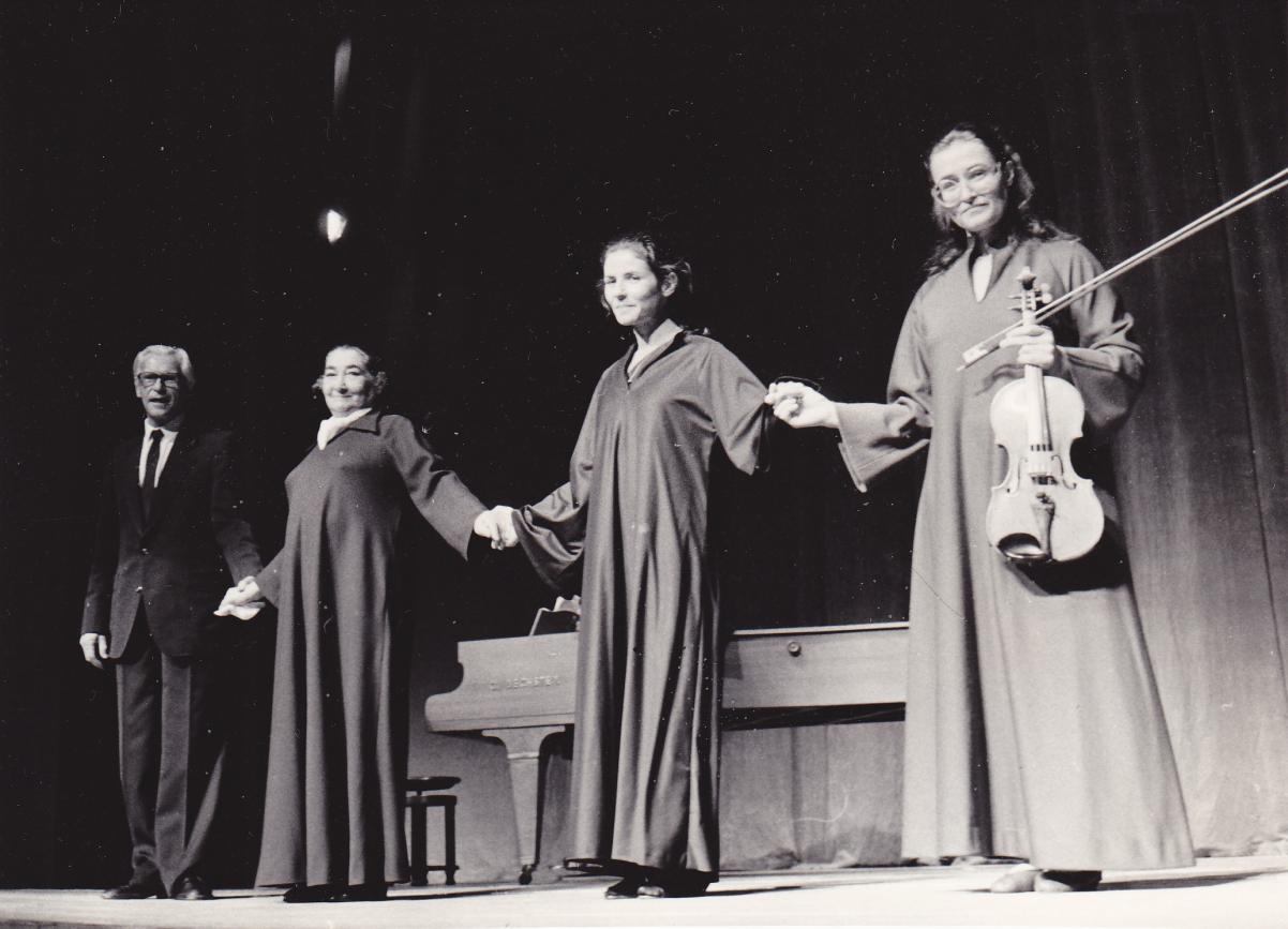 Czarno-biała fotografia. Lin Jaldati Ensemble 1984. Jeden starszy mężczyzna w garniturze i trzy kobiety w długich szatach stoją na scenie, trzymając się za ręce. Kobieta, która stoi z prawej strony, trzyma skrzypce.