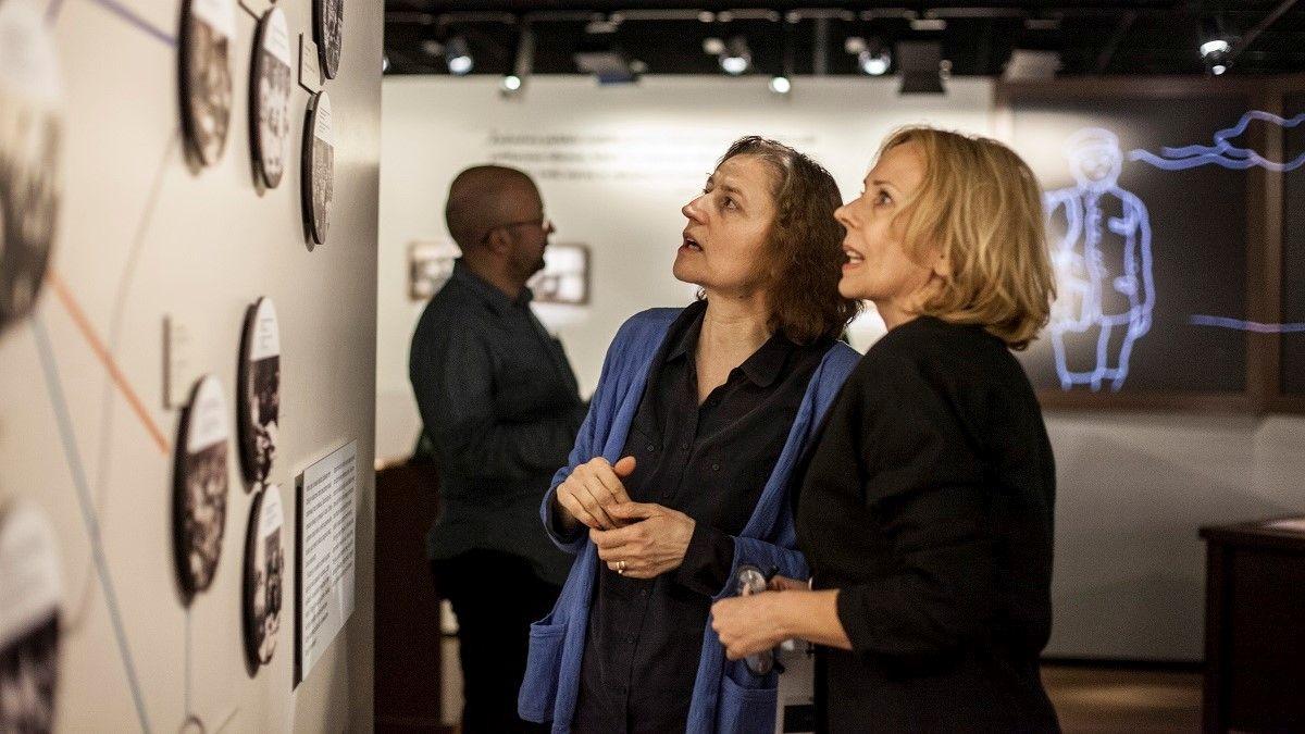 Dwie dorosłe kobiety stoją przed ścianą z ekspozycji, w zaciemnionym pomieszczeniu