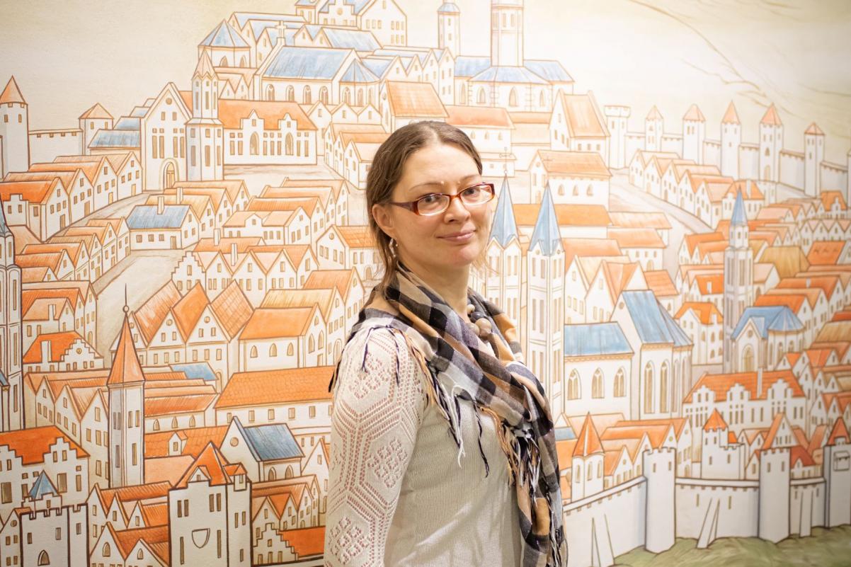 Alla Marchenko - brunetka w okularach i chustce na szyi - stoi przy ścianie z rysunkiem przedstawiającym miasteczko.