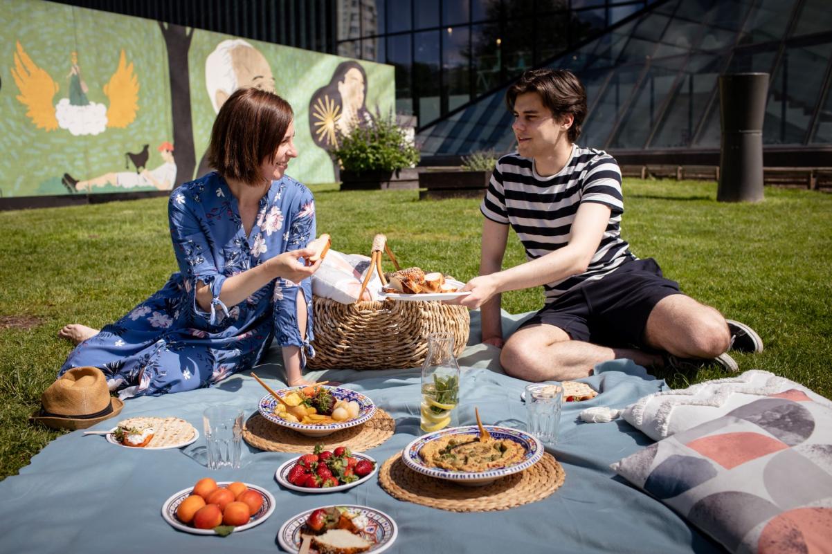 Kobieta i mężczyzna piknikują na łące przed Muzeum POLIN. Na trawie leży koc, na którym znajdują się miski z jedzeniem.