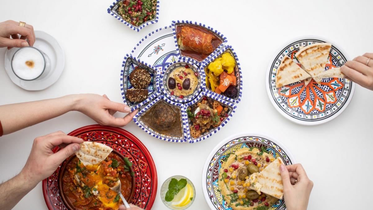 Na stole znajdują się talerze z mezze izraelskimi - pitą, hummusem, falafelami, tahini - i miska z szakszuką. Częstuje się nimi kilka osób - nad talerzami wyciągnięte dłonie.