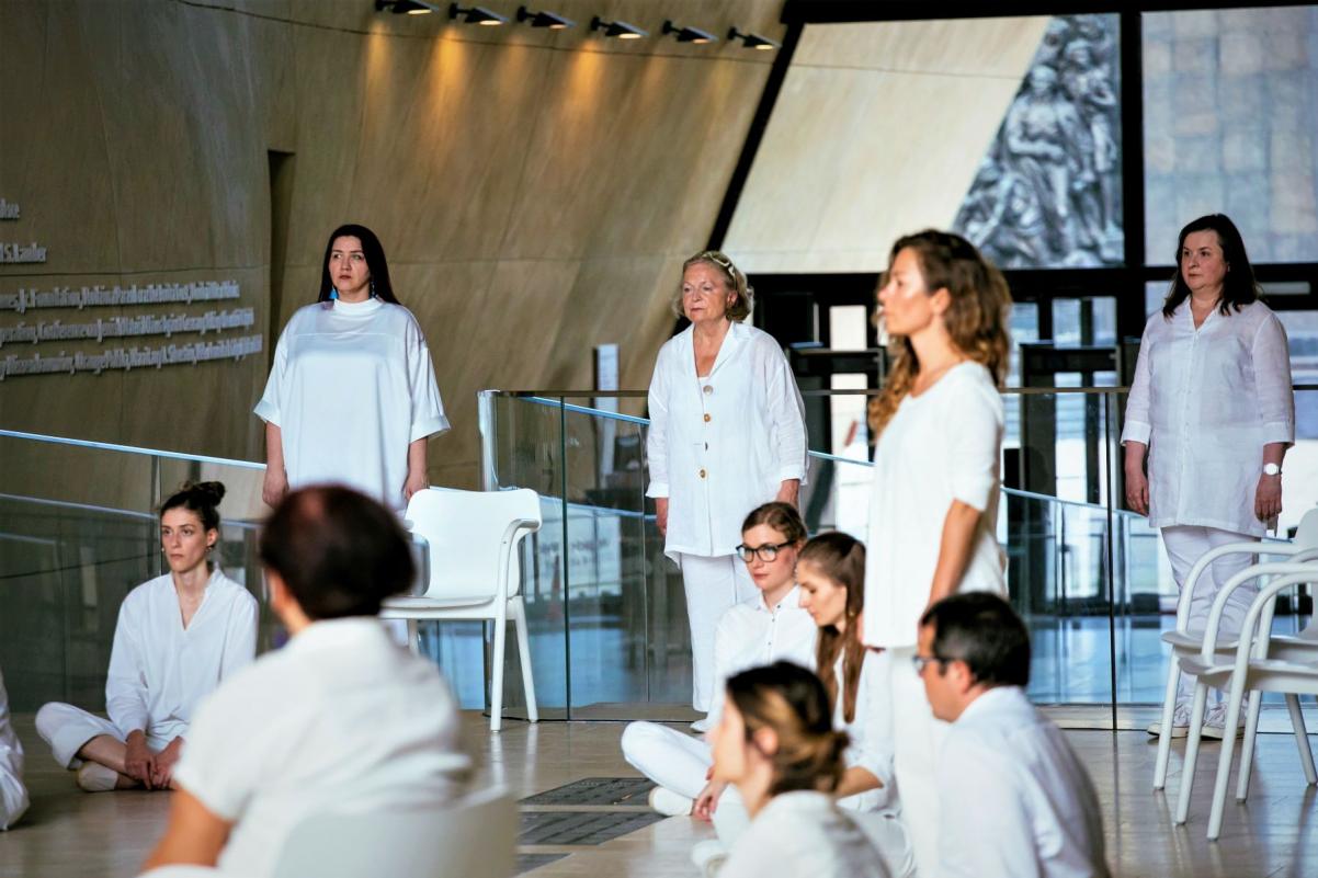 Członkinie chóru POLIN stoją lub siedzą w holu głównym Muzeum POLIN podczas występu.
