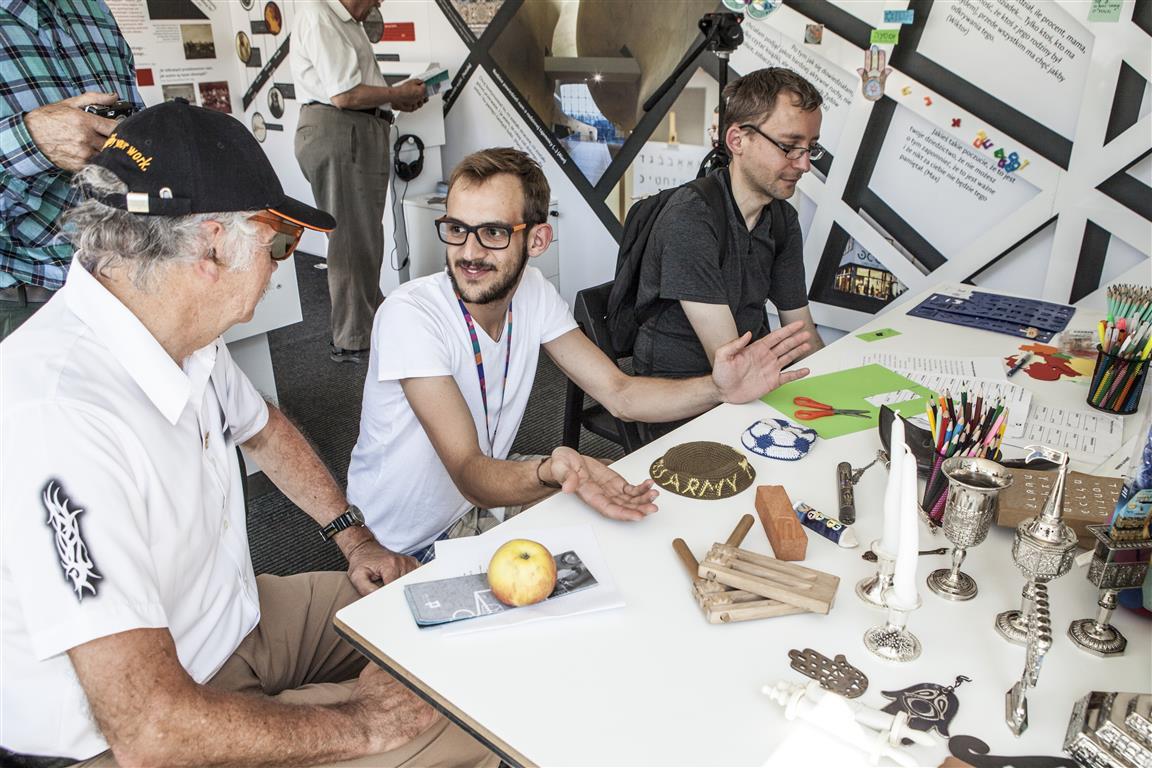 Warsztaty towarzyszące wystawie "Muzeum na kółkach". Trzej mężczyźni siedzą przy stoliku, na którym znajdują się kredki, kolorowe kartki, nożyczki, srebrne besaminki.