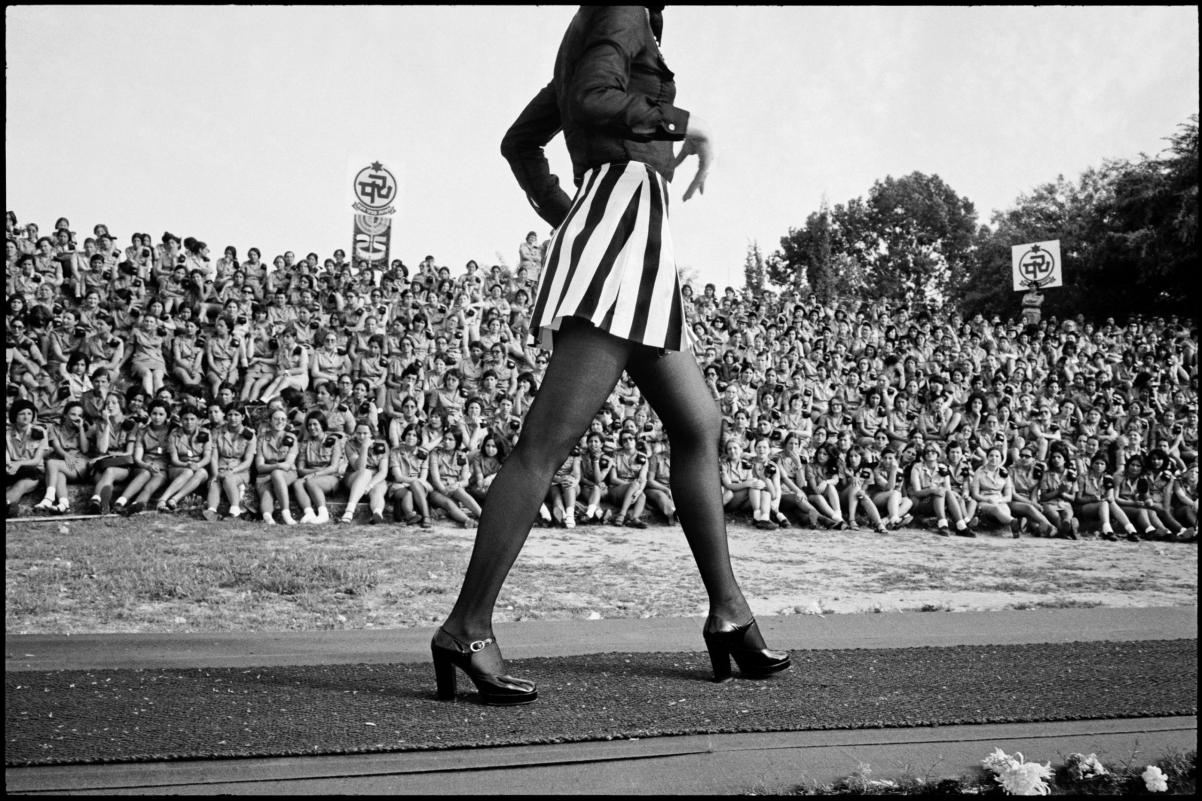 Kadr z filmu "1341 fotosów o miłości i wojnie". Jakaś wysoka kobieta w obcasach idzie po bieżni. Obserwuje ją licznie zgromadzona publiczność na stadionie.