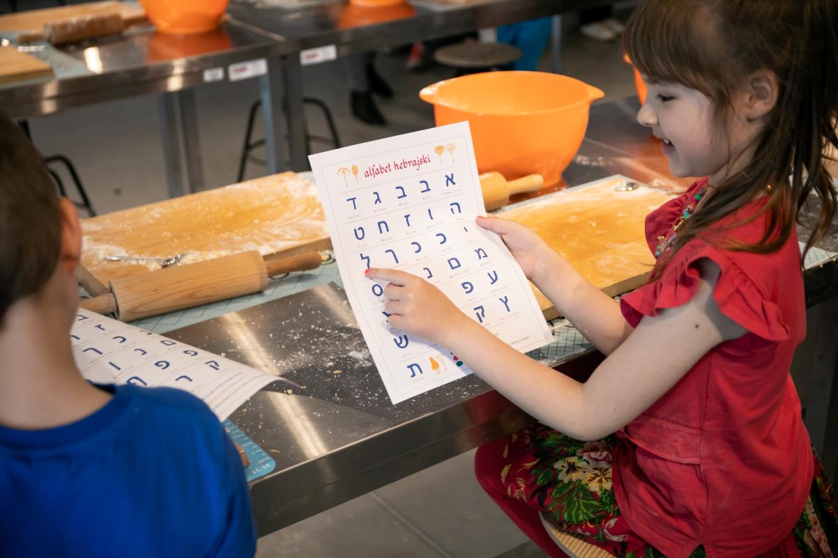 Dziewczynka trzyma kartę z alfabetem hebrajskim i wskazuje na jedną z liter. Obok siedzi chłopiec.