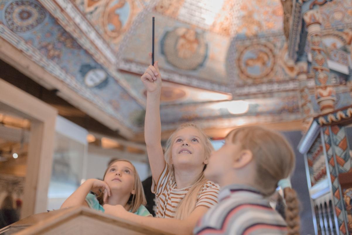 Trzy dziewczynki patrzą na sklepienie synagogi w Gwoźdźcu zrekonstruowane na wystawie stałej w POLIN. Jedna z dziewczynek trzyma ołówek w uniesionej ręce.