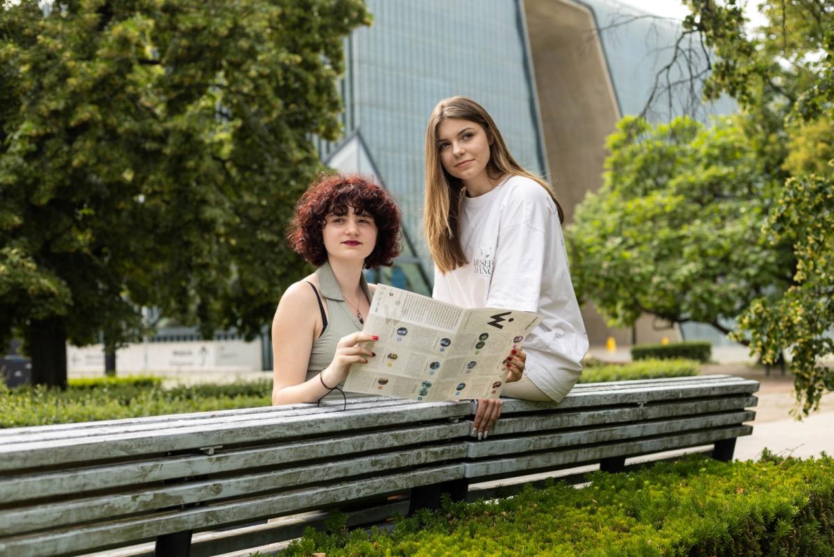 Dwie dziewczyny siedzą na ławce na zielonym skwerze przy budynku Muzeum POLIN. Jedna z nich trzyma broszurę.