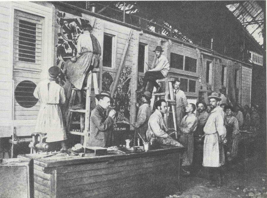 Czarno-białe zdjęcie przedstawia grupę osób przy wagonie kolejowym. Niektórzy stoją na drabinie i malują wagon.