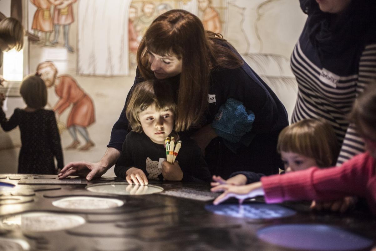 Warsztaty dla szkół w Muzeum POLIN. Na zdjęciu dzieci z opiekunami pochylają się z zainteresowaniem nad stołem z podświetlonymi okienkami.