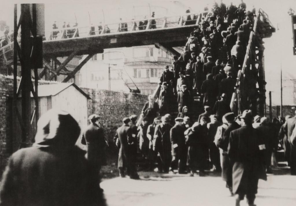 Warsztaty edukacyjne dla szkół ponadpodstawowych. Na zdjęciu archiwalnym z czasów istnienia getta warszawskiego widać grupy ludzi w czarnych ubraniach, którzy przechodzą kładką nad murem getta