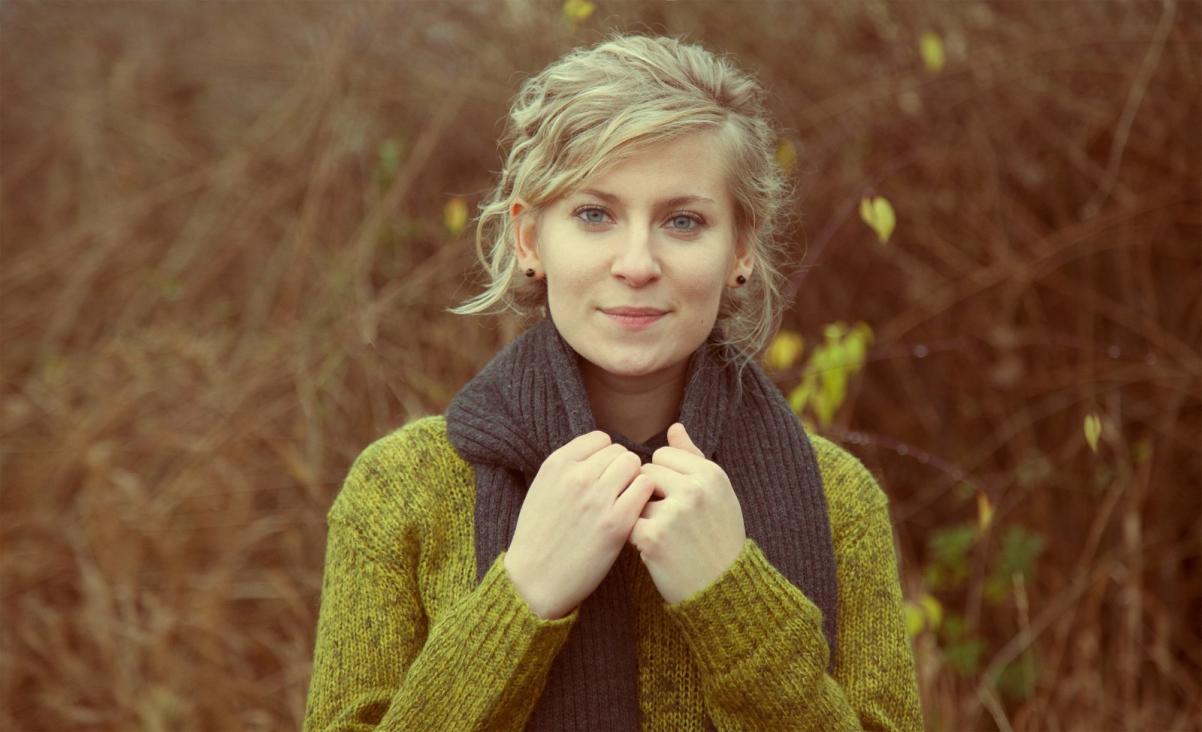 Zdjęcie portretowe przedstawia blondynkę w zielonym swetrze i czarnym szalikiem na szyi.