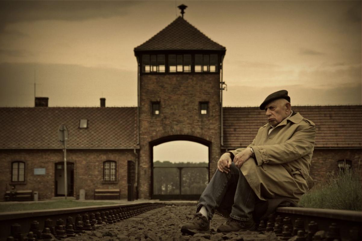 Starszy człowiek siedzi przed bramą obozu koncentracyjnego.