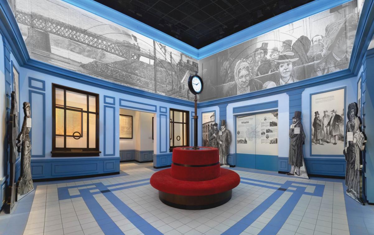 Warsztaty edukacyjne dla szkół ponadpodstawowych. Na zdjęciu: sala dworca, zrekonstruowana w przestrzeni muzeum. Na środku pomieszczenia czerwone okrągłe siedzisko, w którym na środku tkwi na rurze wielki zegar dworcowy