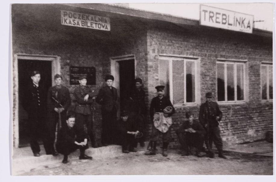Czarno-białe zdjęcia przedstawia grupę ciepło ubranych ludzi przed budynkiem dawnego dworca kolejowego w Treblince