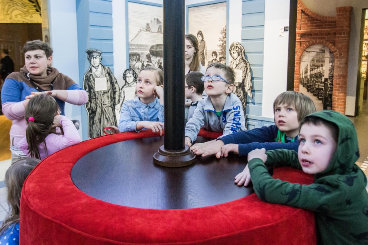 Grupa dzieci stoi wokół czarno-czerwonego koła, z czarną rurą wychodzącą ze środka koła. Niektóre patrzą w górę. Słuchają, co mówi kobieta po lewej stronie zdjęcia.