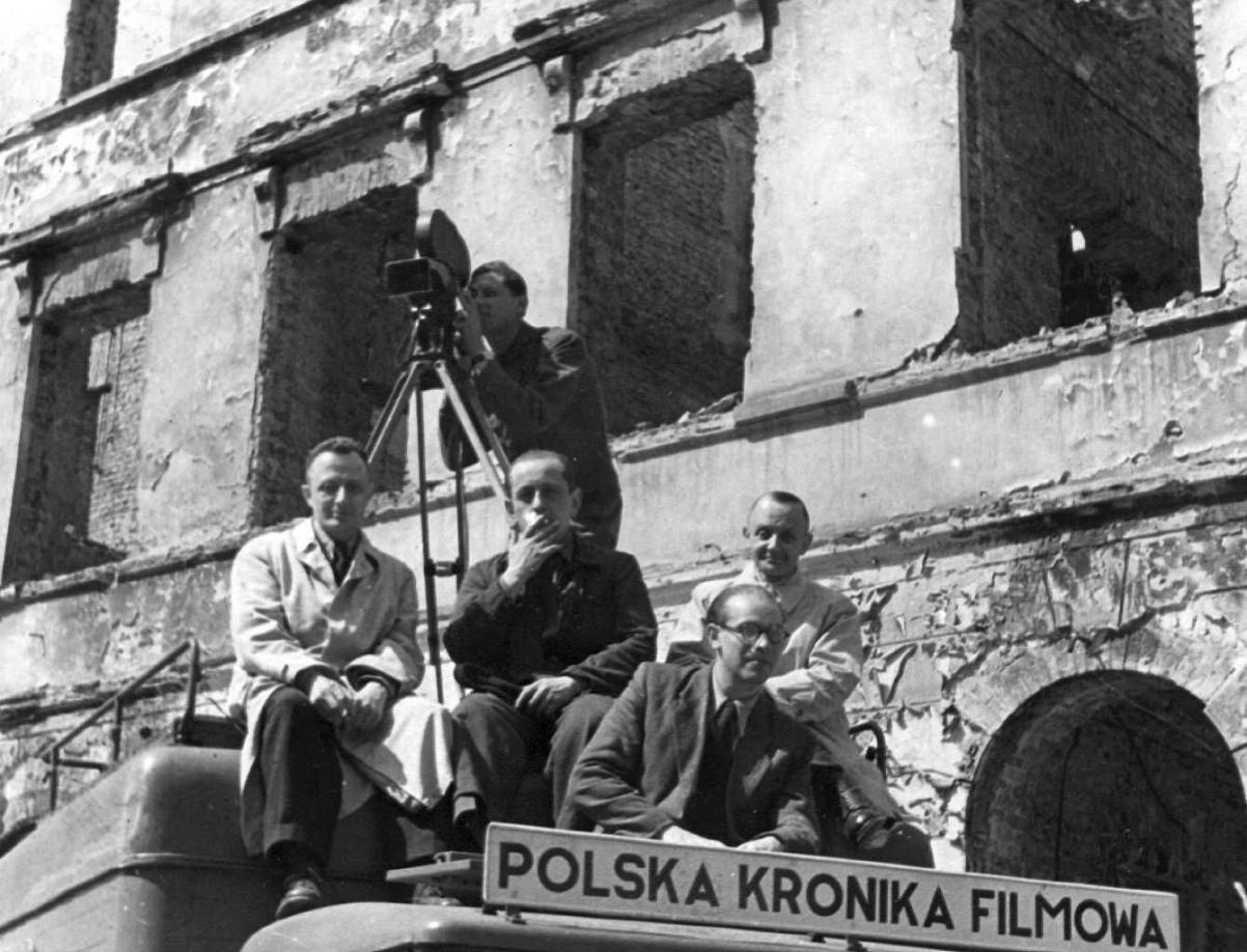 Grupa mężczyzn siedzi na dachu, jeden stoi przy kamerze. Widać szyld Polska Kronika Filmowa.