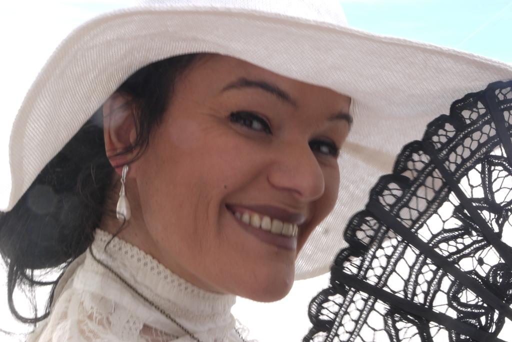 Brunetka w białym kapeluszu uśmiecha się, trzymając w dłoni czarny koronkowy wachlarz.