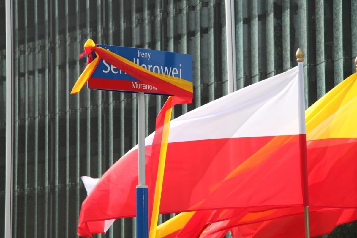 Na zdjęciu niebiesko-czerwona tabliczka z nazwą ulicy: ul. Ireny Sendlerowej, w tle flaga polski, oraz żółto-czerwona flaga Warszawy.