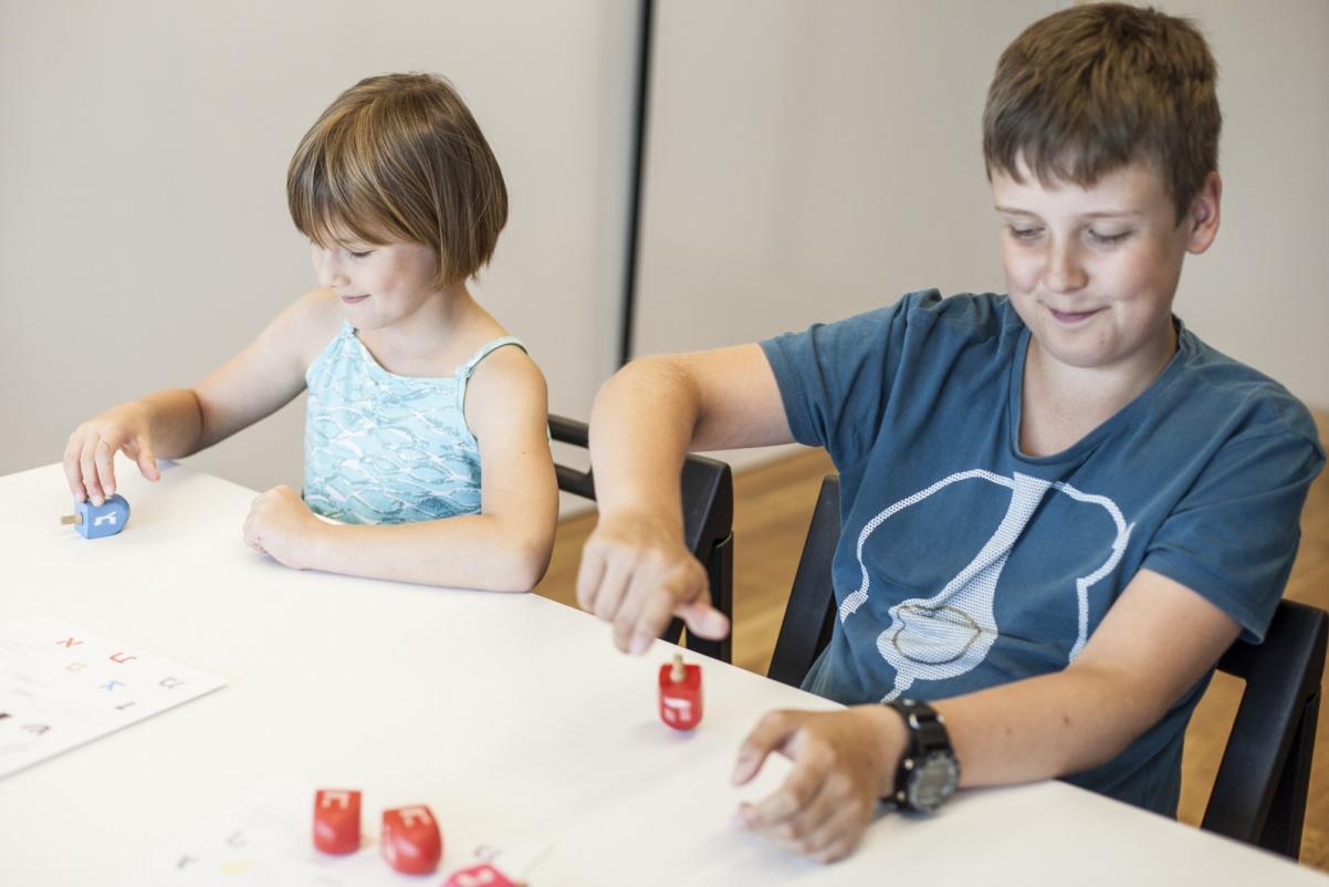 \Chłopiec i dziewczynka siedzą przy białym stole. Przed nimi małe drewniane zabawki - małe bączki zwane drejdlami. Dzieci bawią się nimi.