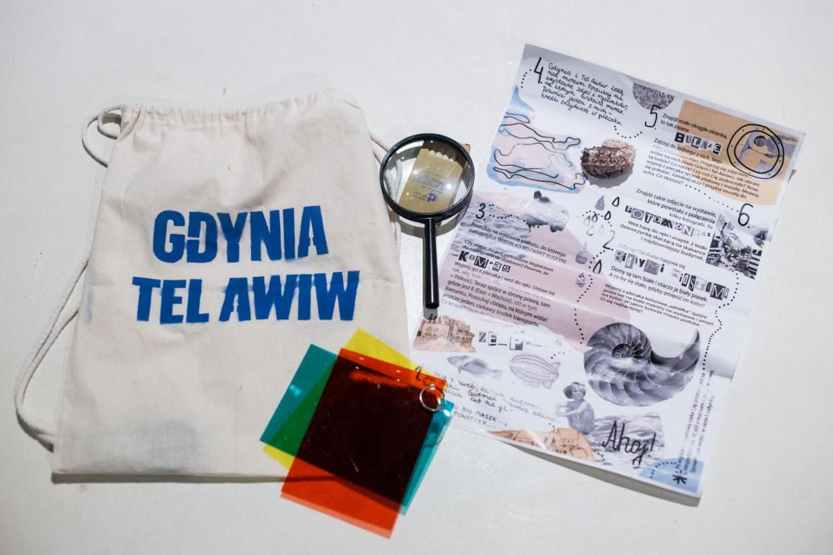 Rodzinna ścieżka zwiedzania wystawy "Gdynia - Tel Awiw" w Muzeum POLIN. Na zdjęciu: Plecak podróżnika: biały plecak-worek z niebieskim napisem "Gdynia - Tel Awiw", kolorowe folie, lupa, plan detektywa