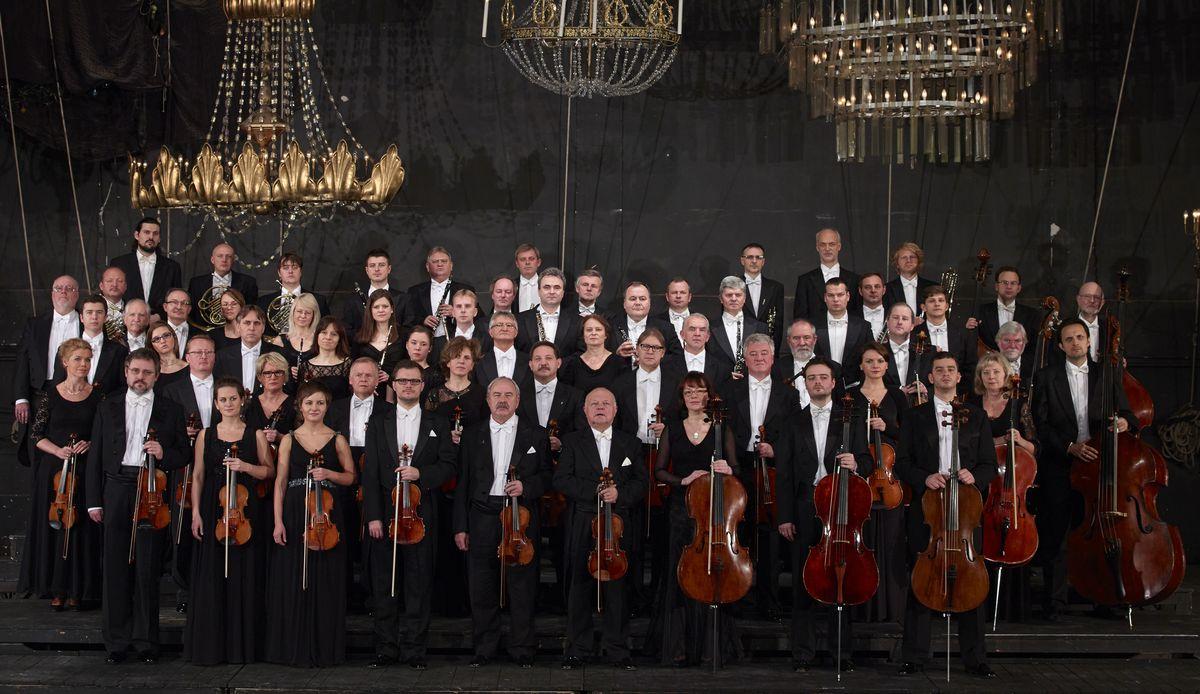 Członkowie orkiestry Sinfonia Varsovia, z instrumentami w dłoniach. W sali piękne żyrandole.
