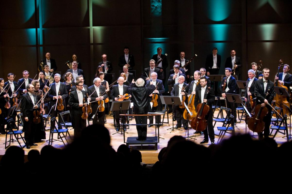 Scena lekko oświetlona, na której stoją z instrumentami członkowie orkiestry wraz z dyrygentem w czasie występu.