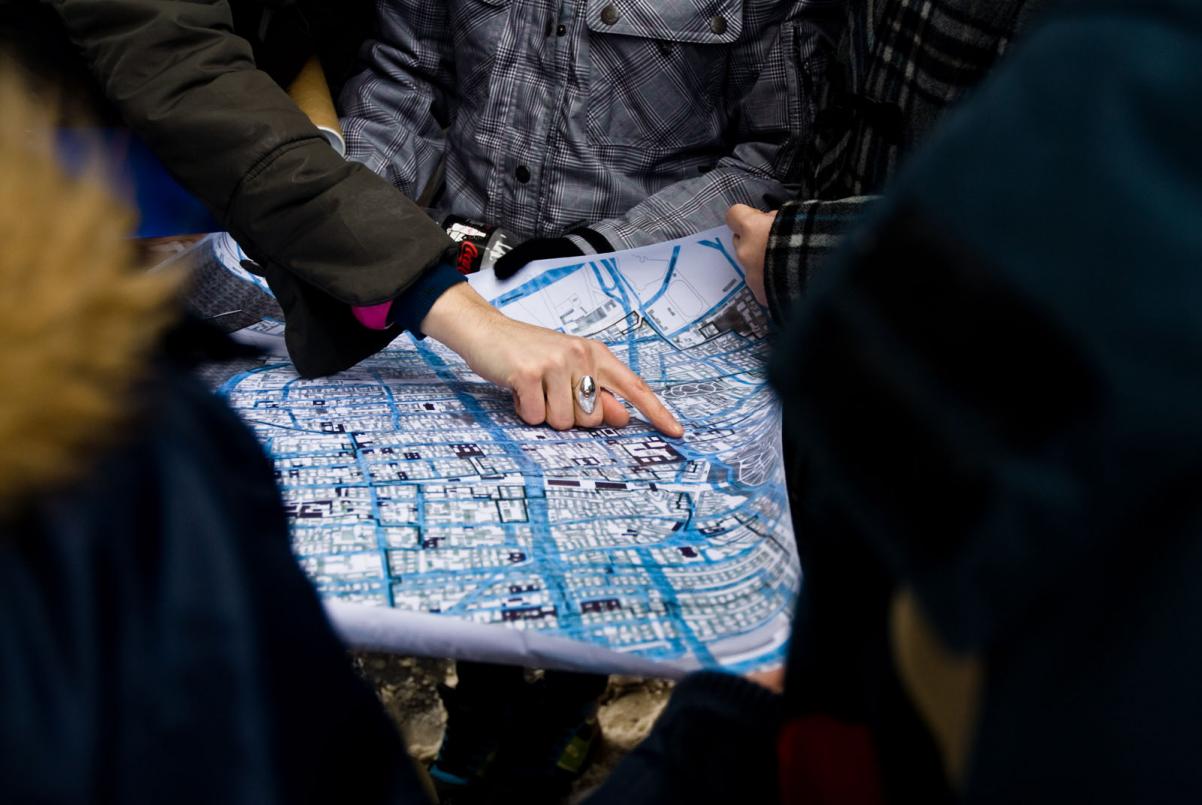 Wędrujący Uniwersytet Muranowski - spotkanie XIII. Na zdjęciu: widok mapy Muranowa, trzymanej przez kilka osób. Widać dłoń jednej z nich, wskazującej jakiś punkt na mapie.